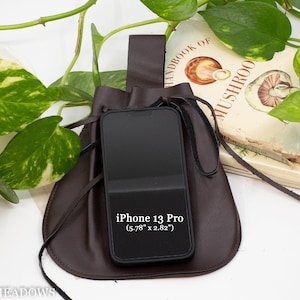 A brown leather pouch with a drawstring closure and a leather strap. The pouch is holding a black iPhone. The pouch is sitting on a white surface with green leafy plants in the background.