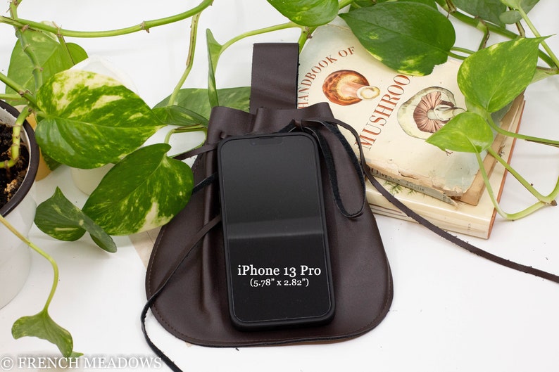A brown leather pouch with a drawstring closure and a leather strap. The pouch is holding a black iPhone. The pouch is sitting on a white surface with green leafy plants in the background.