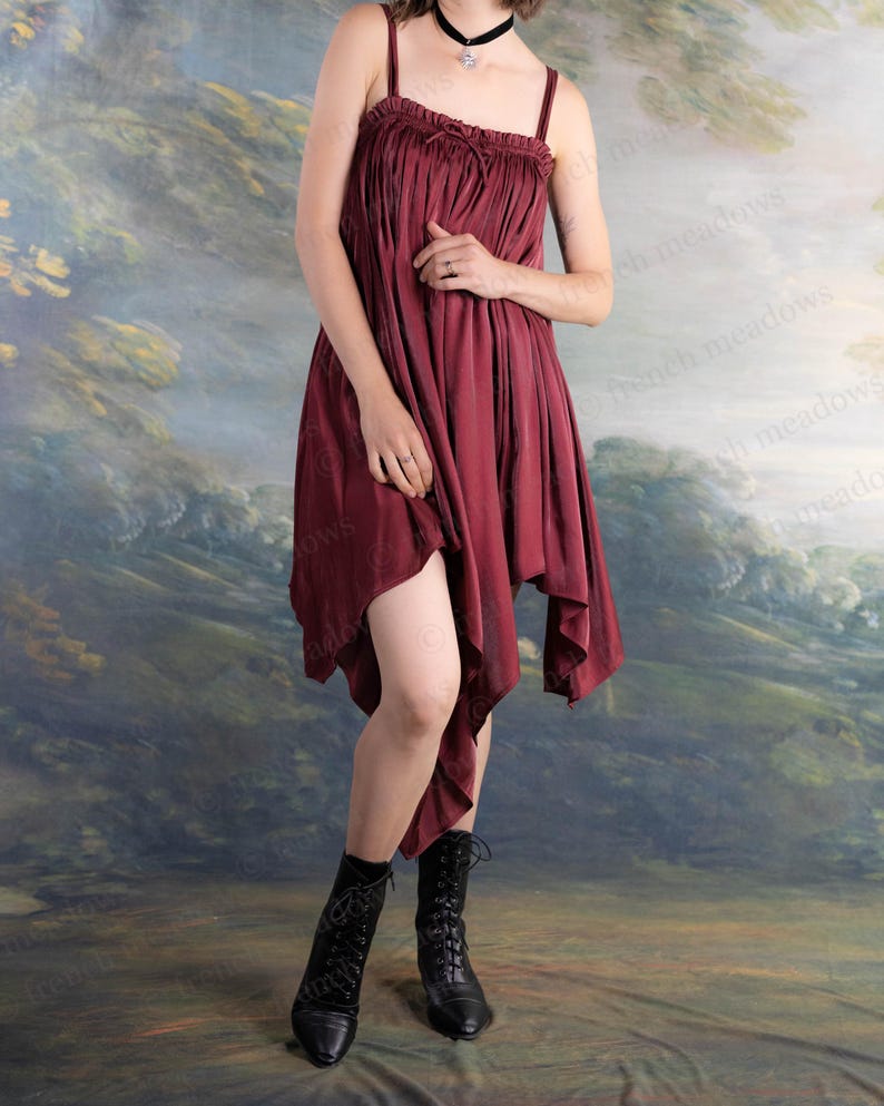 May include: A burgundy-colored dress with spaghetti straps and a gathered bodice. The dress has an asymmetrical hemline and is paired with black lace-up boots. The background features a landscape painting.