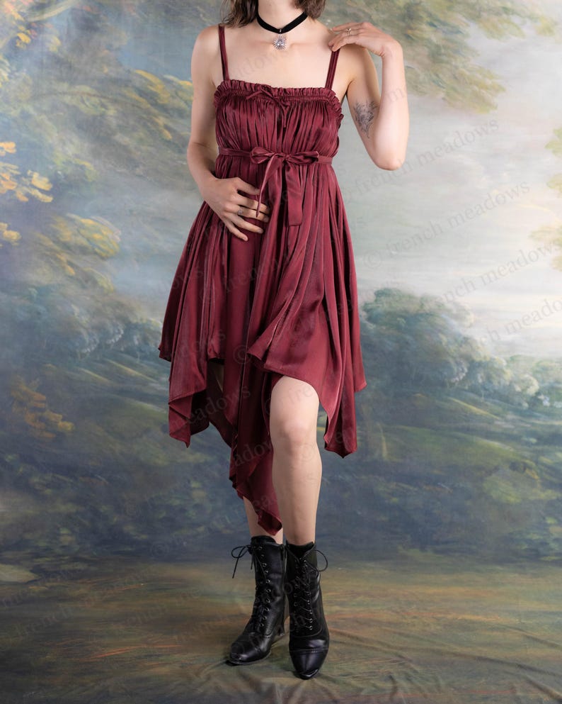 May include: A burgundy-colored dress with spaghetti straps and a flowing, asymmetrical hemline. The dress features a cinched waist with a matching tie and is paired with black lace-up boots. The background is a painted landscape.