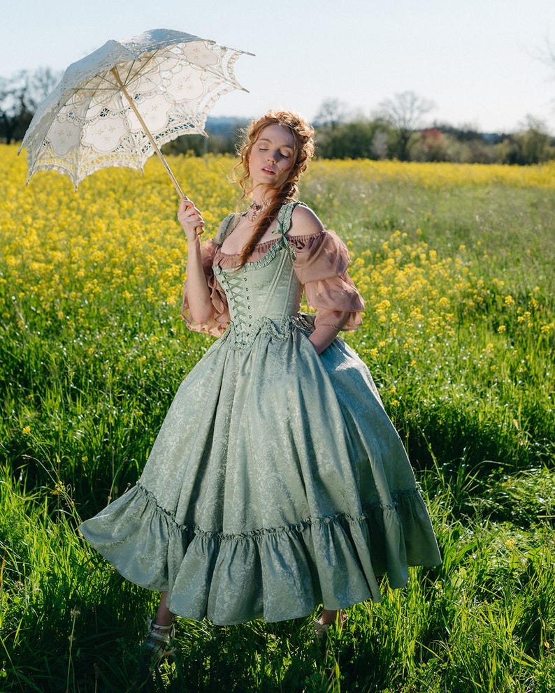 Puede incluir: Un vestido verde claro de estilo vintage con corpi&ntilde;o de cors&eacute; y falda con volantes. El vestido presenta un estampado floral y se combina con un parasol de encaje color crema. Las mangas son de color rosa p&aacute;lido. El fondo es un campo de flores amarillas.