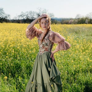 May include: A woman in a field of yellow flowers wears a vintage-style dress. The dress features a floral patterned bodice, olive green skirt, and sheer, puffed sleeves. The model is standing in a field of yellow flowers.