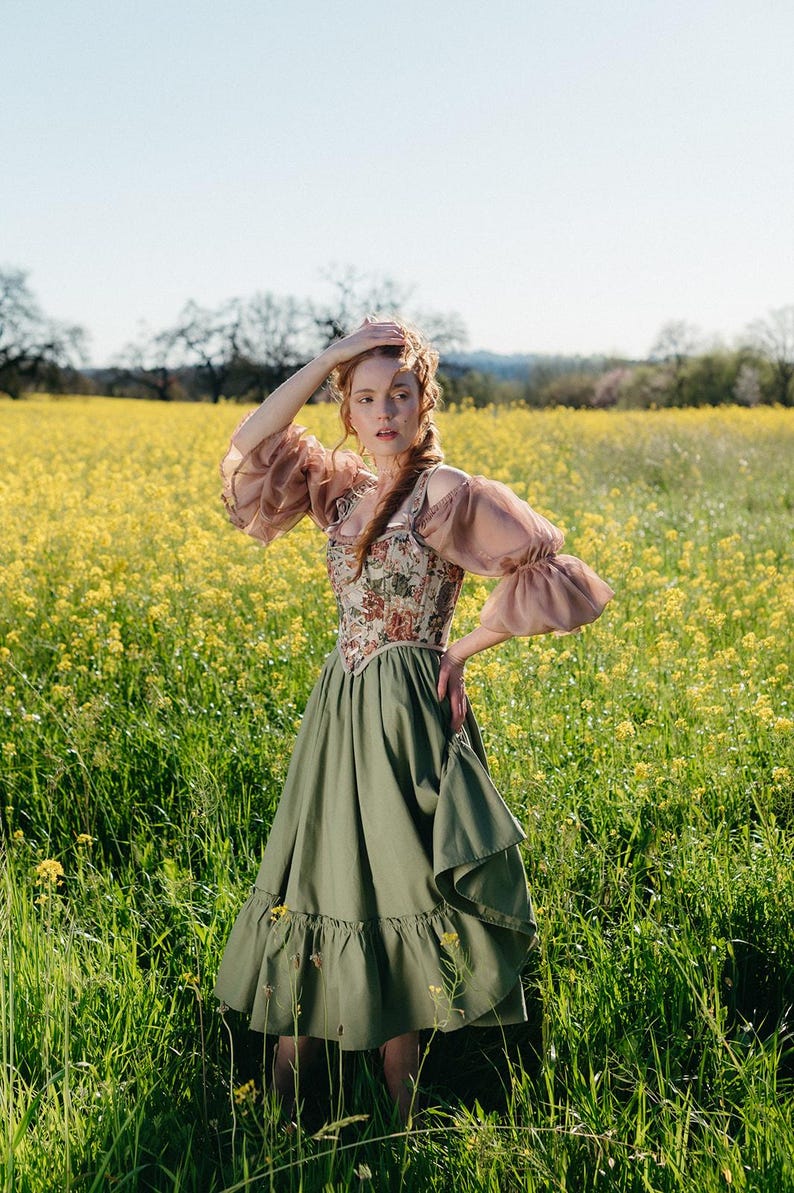 May include: A woman in a field of yellow flowers wears a vintage-style dress. The dress features a floral patterned bodice, olive green skirt, and sheer, puffed sleeves. The model is standing in a field of yellow flowers.