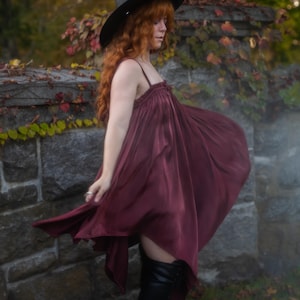 May include: A burgundy-colored dress with spaghetti straps and a flowing skirt. The dress is paired with a black witch hat and black knee-high boots. The background features a stone wall with ivy and autumn foliage.