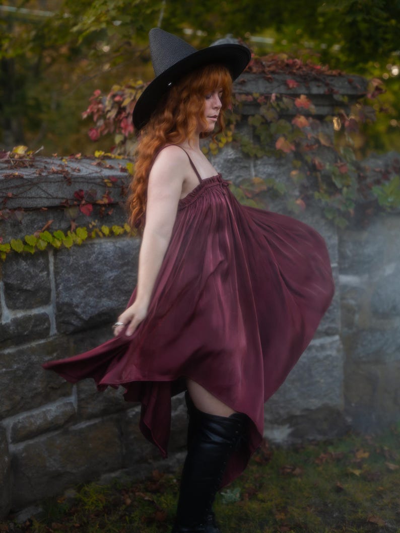 May include: A burgundy-colored dress with spaghetti straps and a flowing skirt. The dress is paired with a black witch hat and black knee-high boots. The background features a stone wall with ivy and autumn foliage.