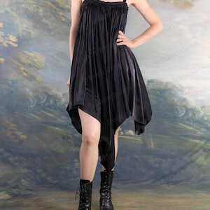 Puede incluir: Vestido negro asim&eacute;trico con tirantes finos y escote con volantes. El vestido tiene un dise&ntilde;o fluido y drapeado y se combina con botas negras con cordones. El fondo presenta un paisaje pintado con colores apagados.