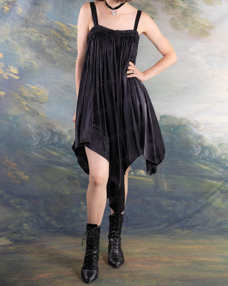 Puede incluir: Vestido negro asim&eacute;trico con tirantes finos y escote con volantes. El vestido tiene un dise&ntilde;o fluido y drapeado y se combina con botas negras con cordones. El fondo presenta un paisaje pintado con colores apagados.