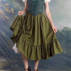 Puede incluir: Un vestido verde y blanco con un corpiño estilo corsé y una falda con volantes. El corpiño es de un verde más oscuro con cordones y correas verdes. La falda es de un verde más claro y las mangas son blancas con ribete de encaje. El vestido se combina con zapatos marrones con cordones.