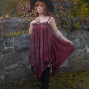 May include: A burgundy dress with spaghetti straps and a flowing, asymmetrical hemline. The dress is paired with black knee-high boots and a wide-brimmed black hat. The background features a stone wall and autumn foliage.