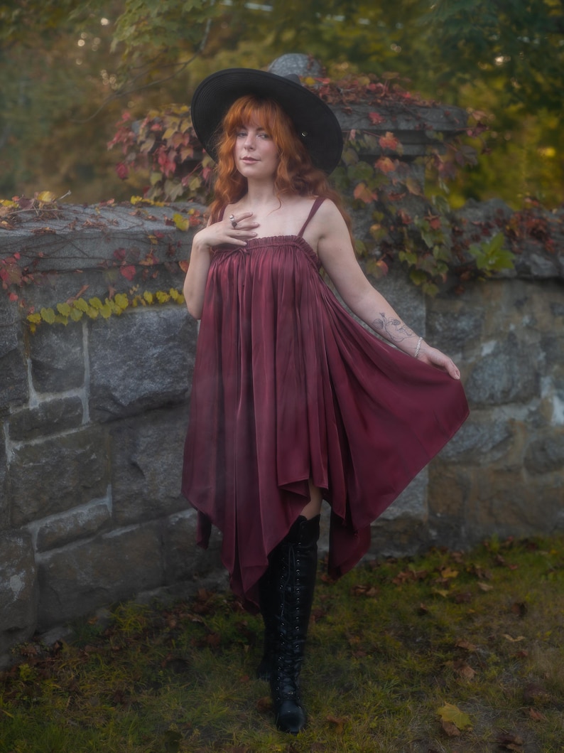 May include: A burgundy dress with spaghetti straps and a flowing, asymmetrical hemline. The dress is paired with black knee-high boots and a wide-brimmed black hat. The background features a stone wall and autumn foliage.
