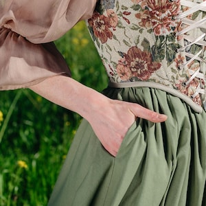 May include: A floral tapestry corset with white lacing is paired with a sage green skirt. The outfit includes a sheer, blush-colored sleeve. The person's hand is in the skirt pocket.