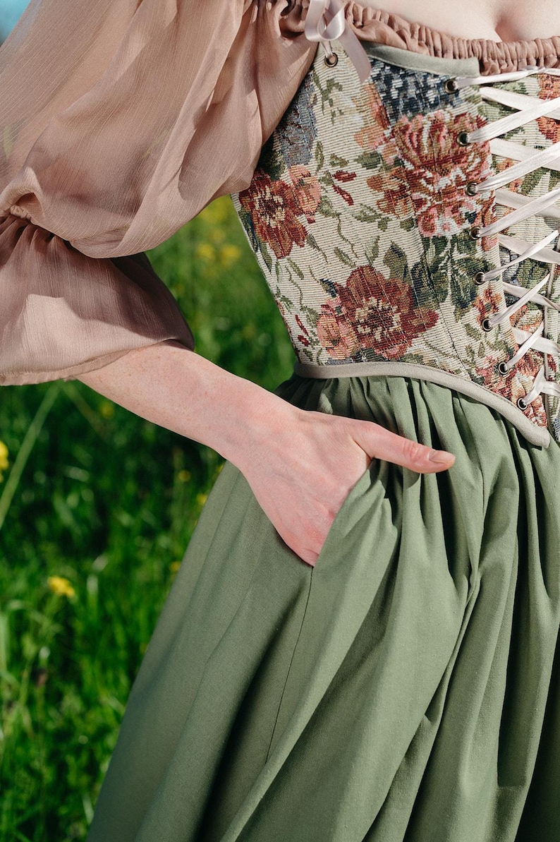 May include: A floral tapestry corset with white lacing is paired with a sage green skirt. The outfit includes a sheer, blush-colored sleeve. The person's hand is in the skirt pocket.