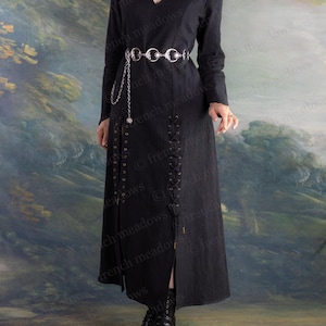 May include: A long black dress with a V-neck and long sleeves. The dress features a silver belt with chain detailing and a slit up the front. The dress is paired with black boots. The background is a painted landscape.