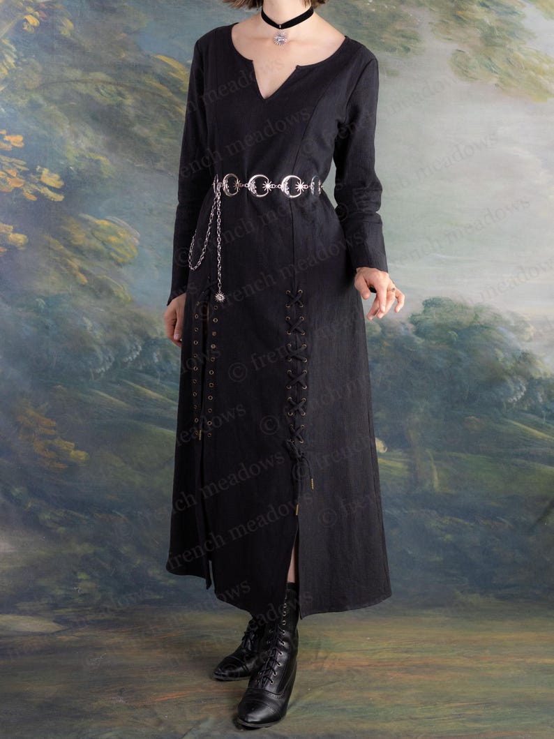 May include: A long black dress with a V-neck and long sleeves. The dress features a silver belt with chain detailing and a slit up the front. The dress is paired with black boots. The background is a painted landscape.