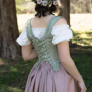 Puede incluir: Un cors&eacute; verde claro con un estampado floral y espalda con cordones. Se combina con una blusa blanca con mangas abullonadas y una falda larga rosa empolvado. Una corona de flores adorna la cabeza.