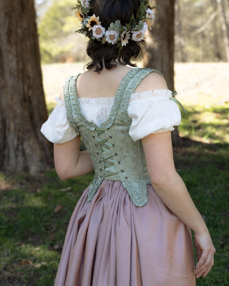 Puede incluir: Un cors&eacute; verde claro con un estampado floral y espalda con cordones. Se combina con una blusa blanca con mangas abullonadas y una falda larga rosa empolvado. Una corona de flores adorna la cabeza.