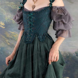 May include: A dark teal renaissance-style dress with a lace-up bodice and off-the-shoulder sleeves. The dress features a layered skirt and gray, ruffled sleeves. Green ribbon accents the straps. A gold pendant necklace is visible.