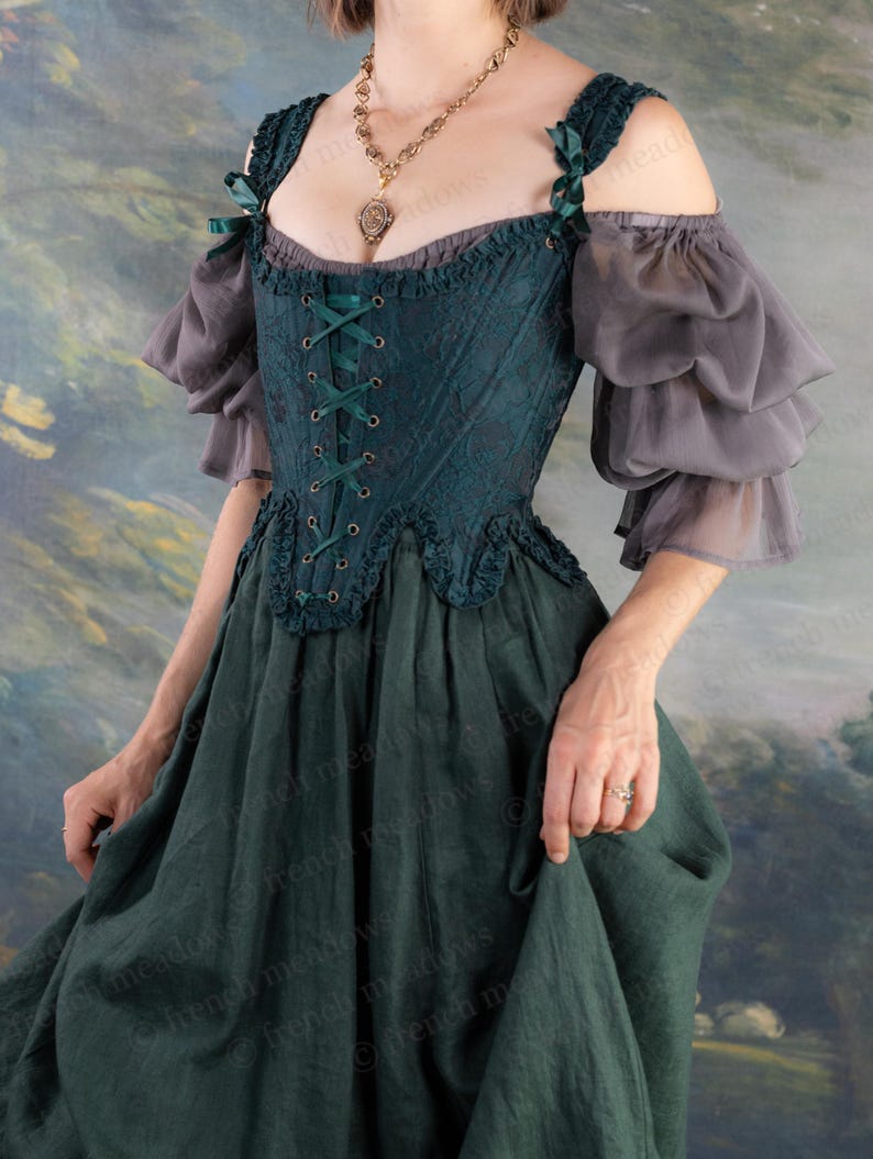 May include: A dark teal renaissance-style dress with a lace-up bodice and off-the-shoulder sleeves. The dress features a layered skirt and gray, ruffled sleeves. Green ribbon accents the straps. A gold pendant necklace is visible.