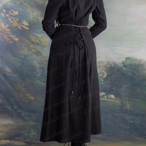 May include: A long black dress with long sleeves and a laced back. The dress features a round neckline and a silver chain belt. The hemline falls to the ankles. The background is a painted landscape with trees and a sky.