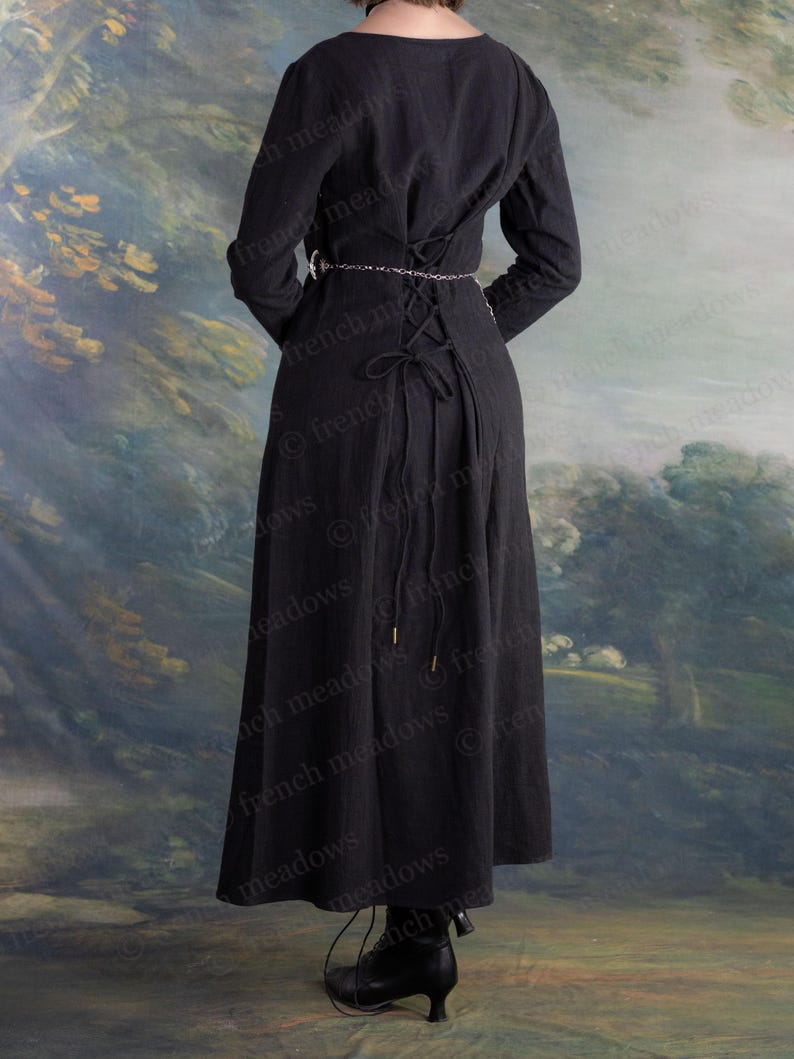 May include: A long black dress with long sleeves and a laced back. The dress features a round neckline and a silver chain belt. The hemline falls to the ankles. The background is a painted landscape with trees and a sky.