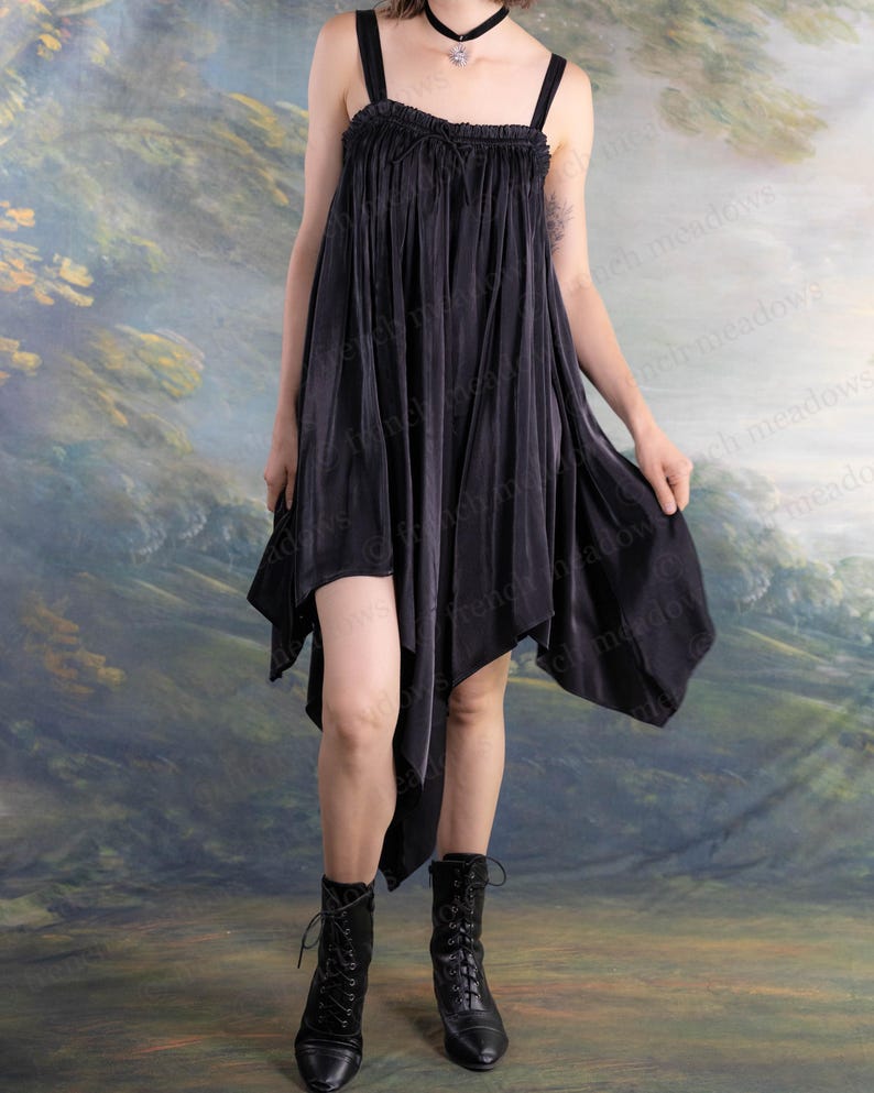 Puede incluir: Vestido negro asim&eacute;trico con tirantes finos y escote con volantes. El vestido tiene un dobladillo fluido en forma de pa&ntilde;uelo y se combina con botas negras con cordones. El fondo es un paisaje pintado.
