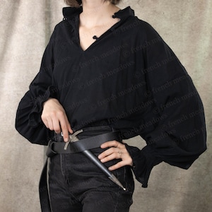 May include: A black, long-sleeved shirt with a ruffled collar and cuffs. The shirt has a V-neckline and is fastened with a series of small buttons. A silver dagger is tucked into a black leather belt worn over black pants.