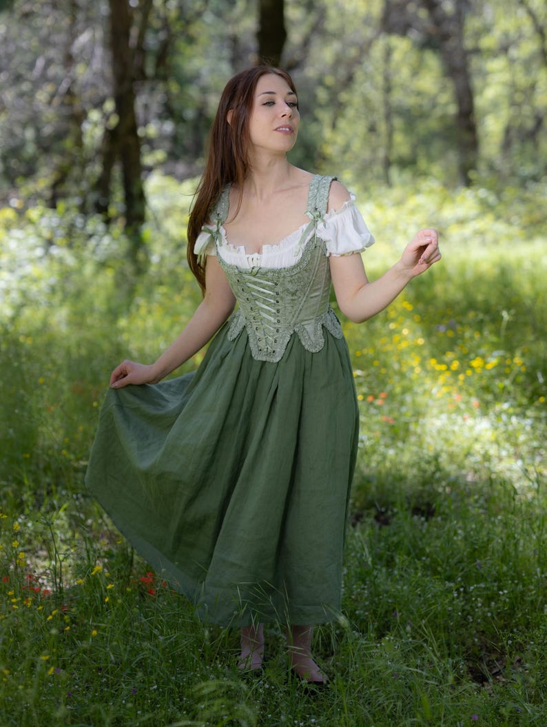 Puede incluir: Un vestido verde y blanco de estilo renacentista. El corpi&ntilde;o es de color verde claro con detalles intrincados y un frente con cordones. La falda es de un verde liso y fluido. Las mangas son cortas y blancas. El vestido se usa al aire libre en una zona herbosa.