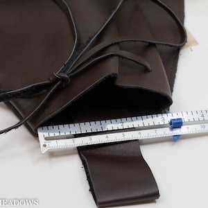 A brown leather pouch with a black leather strap and a metal ruler measuring 12 centimeters. The pouch is open and the strap is tied in a knot. The ruler is placed on top of the pouch and the strap.