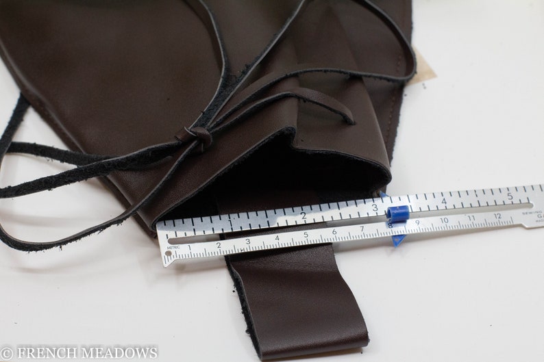 A brown leather pouch with a black leather strap and a metal ruler measuring 12 centimeters. The pouch is open and the strap is tied in a knot. The ruler is placed on top of the pouch and the strap.