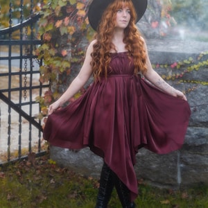 Könnte beinhalten: Ein bordeauxfarbenes Kleid mit fließendem Rock und eng anliegendem Oberteil. Das Kleid wird mit schwarzen kniehohen Stiefeln und einem breitkrempigen schwarzen Hut kombiniert. Die Person auf dem Bild hat lange, rote Haare und steht in einem Garten.