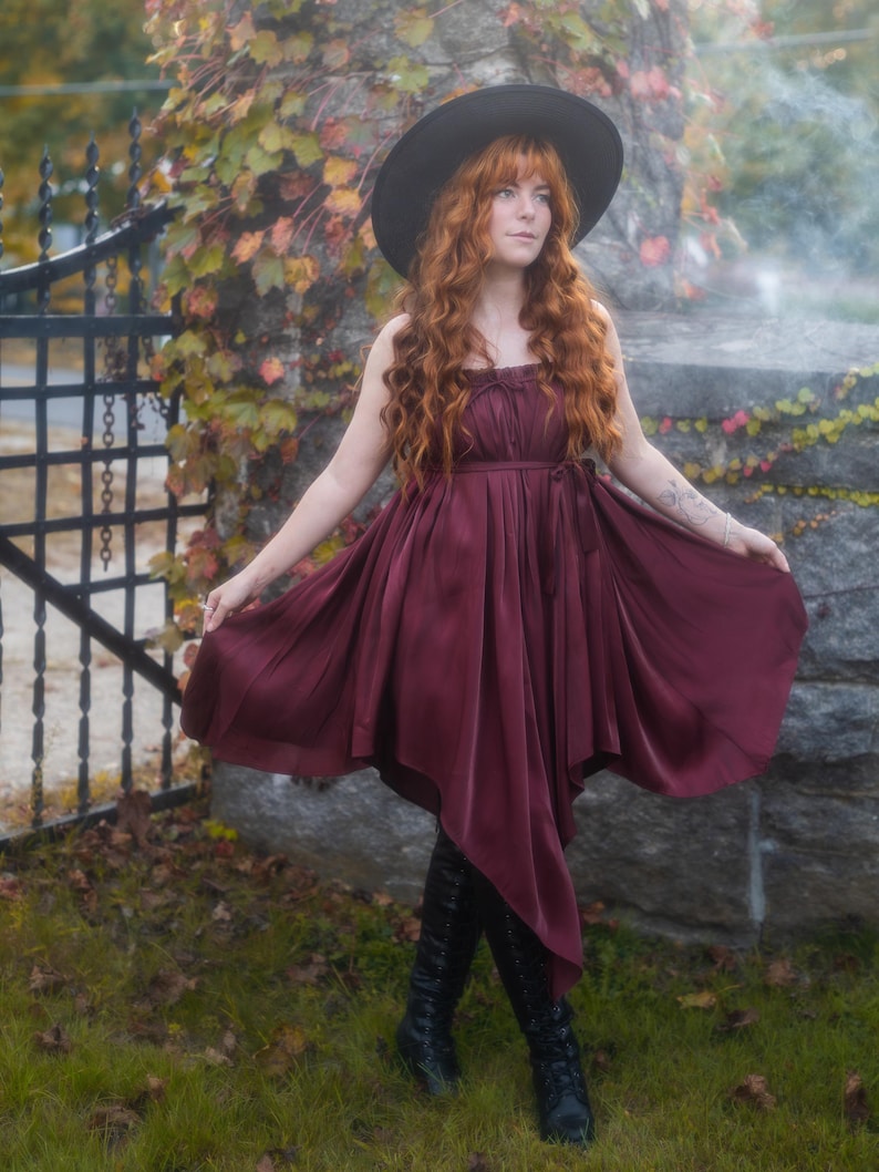 May include: A burgundy dress with a flowing skirt and a fitted bodice. The dress is paired with black knee-high boots and a wide-brimmed black hat. The person in the image has long, red hair and is standing in a garden setting.