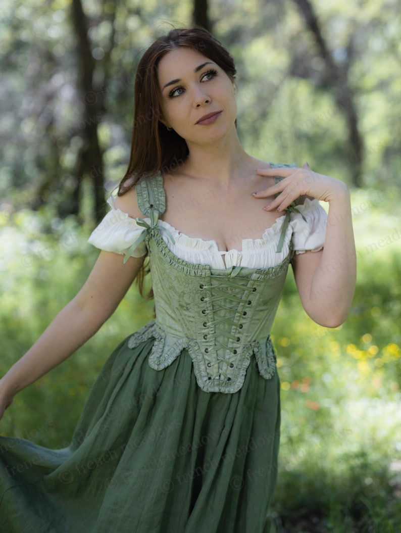 Puede incluir: Un cors&eacute; verde con cordones frontales y bordes con volantes, combinado con una falda verde a juego. La parte superior tiene mangas blancas con lazos de cinta verde. El atuendo recuerda la vestimenta hist&oacute;rica o de fantas&iacute;a.