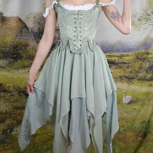 Puede incluir: Vestido cors&eacute; verde salvia con falda en capas. El vestido presenta un corpi&ntilde;o con cordones, mangas descubiertas y adornos delicados. La falda tiene m&uacute;ltiples capas de tela fluida. El vestido recuerda a un estilo de hadas o renacentista.