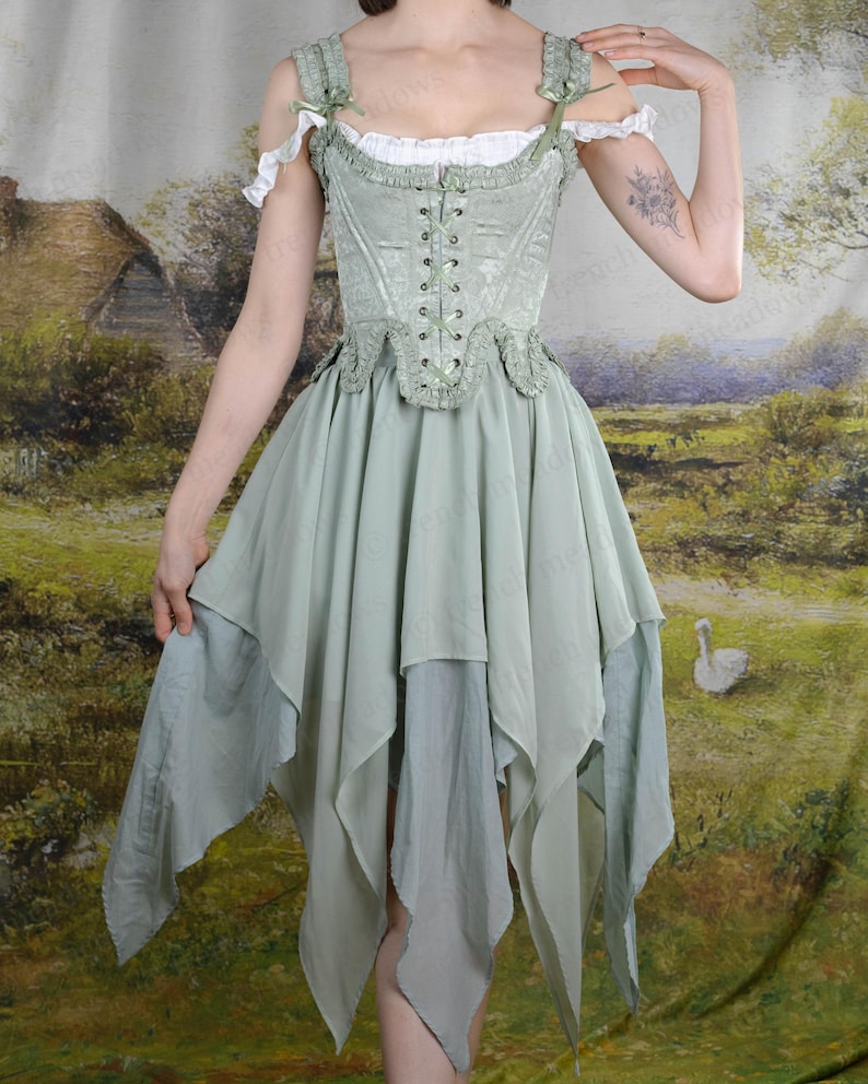 Puede incluir: Vestido cors&eacute; verde salvia con falda en capas. El vestido presenta un corpi&ntilde;o con cordones, mangas descubiertas y adornos delicados. La falda tiene m&uacute;ltiples capas de tela fluida. El vestido recuerda a un estilo de hadas o renacentista.