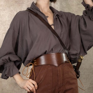 Puede incluir: Una camisa gris de manga larga con un cinturón de cuero marrón y una correa de cuero marrón. La camisa tiene un cuello con volantes y las mangas están fruncidas en las muñecas. El cinturón tiene una hebilla decorativa y se lleva sobre unos pantalones marrones.