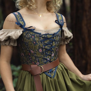 Op de afbeelding: Een korsettop met een donkerblauw en olijfgroen bloemenpatroon, marineblauwe lintveters en schouderstrikken. Het kledingstuk wordt gedragen over een beige off-shoulder blouse met ruches aan de mouwen, gecombineerd met een olijfgroene rok en een bruine riem.