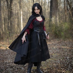 May include: A woman wearing a black hooded cloak, a red and black bodice, a black skirt, and black boots. She is holding a sword in her right hand and is standing in a forest.