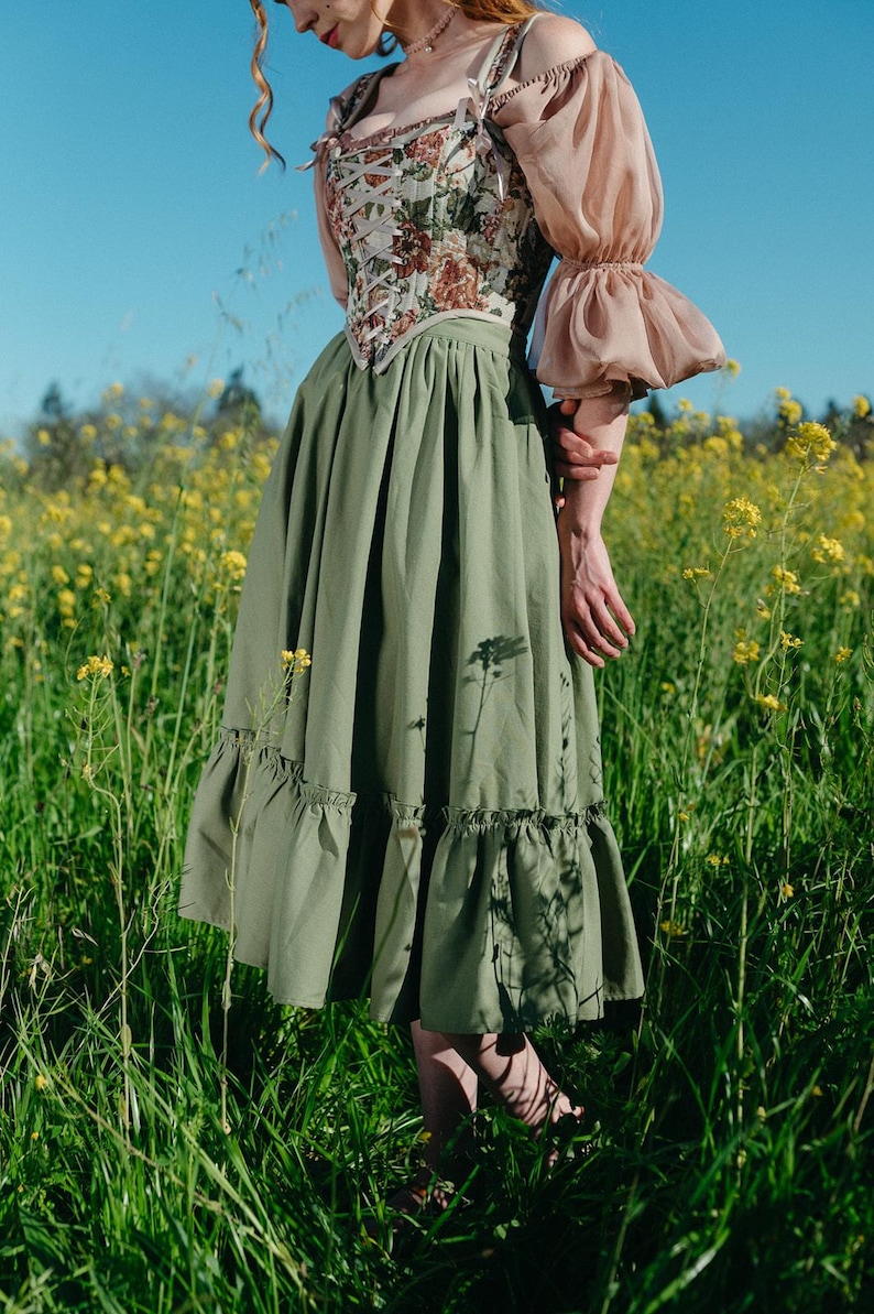 May include: A floral corset top with a green skirt and sheer sleeves. The corset features a delicate floral pattern, and the skirt has a ruffled hem. The sleeves are a light pink color. The outfit is set in a field of yellow flowers.