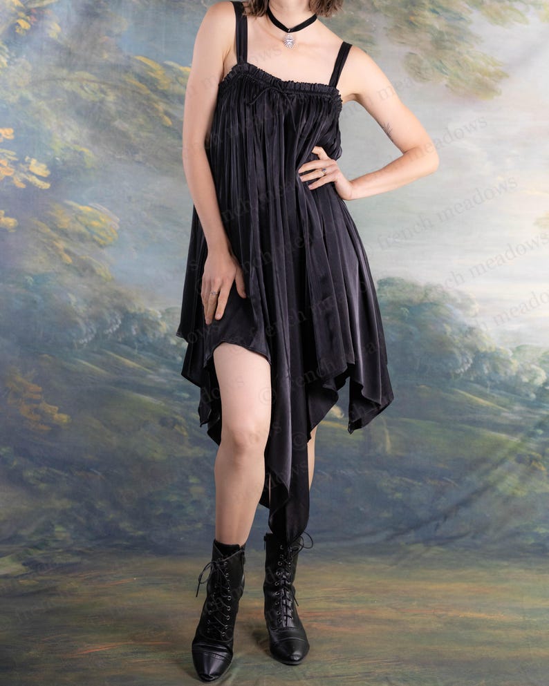 Puede incluir: Un vestido negro asim&eacute;trico con tirantes finos y corpi&ntilde;o con volantes. El vestido tiene un dobladillo fluido en forma de pa&ntilde;uelo y se combina con botas negras con cordones. El fondo presenta un paisaje pintado.