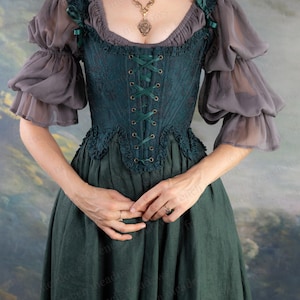 May include: A dark teal corset-style top with a lace-like pattern and green ribbon lacing. The top has a ruffled neckline and is paired with gray, sheer, puffed sleeves. A long, dark green skirt completes the ensemble. A gold pendant necklace is also visible.
