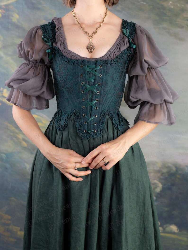 May include: A dark teal corset-style top with a lace-like pattern and green ribbon lacing. The top has a ruffled neckline and is paired with gray, sheer, puffed sleeves. A long, dark green skirt completes the ensemble. A gold pendant necklace is also visible.
