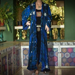 May include: A long, flowing blue robe with white floral accents and a black top. The robe features a tie-dye effect and is adorned with a decorative belt and necklace. The background includes plants and a decorative cabinet.