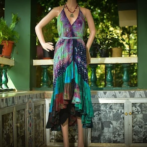 May include: A vibrant tie-dye halter dress displayed on a mannequin. The dress has a V-neck and a flowing, layered skirt in shades of purple, green, blue, and brown. A necklace with a pendant is visible. The background includes greenery and a decorative balcony.