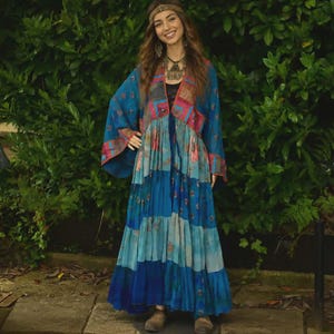 May include: A long, tiered blue dress with a patchwork design on the bodice and sleeves. The dress features shades of blue and teal, with red and pink accents. The person is wearing a headband and jewelry, and is standing in front of a green hedge.