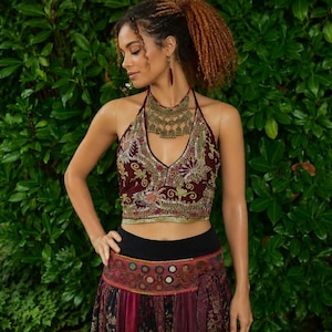 May include: A maroon halter crop top featuring intricate gold and multi-coloured embroidery, paired with a matching maroon and black patterned skirt with circular accents. A large, ornate gold-toned statement necklace rests at the neckline.