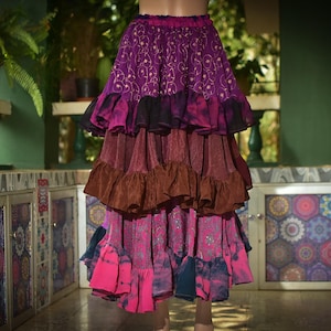 May include: A tiered skirt in shades of purple, brown, and pink. The skirt features a ruffled design and a mix of patterned and solid fabrics. The skirt is designed for a bohemian or festival style.