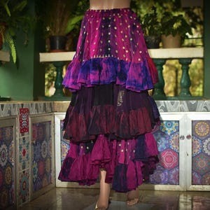 May include: A tiered skirt in shades of pink, purple, and black. The skirt has a flowing, layered design with a mix of solid and patterned fabrics. The skirt appears to be handmade, with a bohemian or festival style.