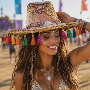Puede incluir: Un sombrero beige con bordados intrincados, lentejuelas y un ala ancha adornada con una hilera de borlas multicolores en tonos rojo, naranja, amarillo, verde, azul y rosa. El sombrero tiene una copa estructurada y un ala plana y ancha.