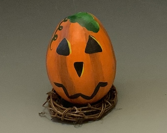 Può includere: Una zucca arancione dipinta a mano con un viso di jack-o'-lantern e uno stelo verde, seduta su un letto di ramoscelli marroni.