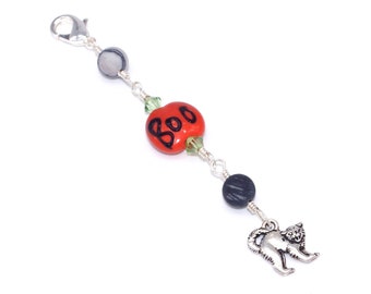 May include: A Halloween-themed charm featuring a red pumpkin bead with "Boo" in black lettering, connected by silver chain to gray and black beads, and a silver cat charm. The charm has a silver clasp.
